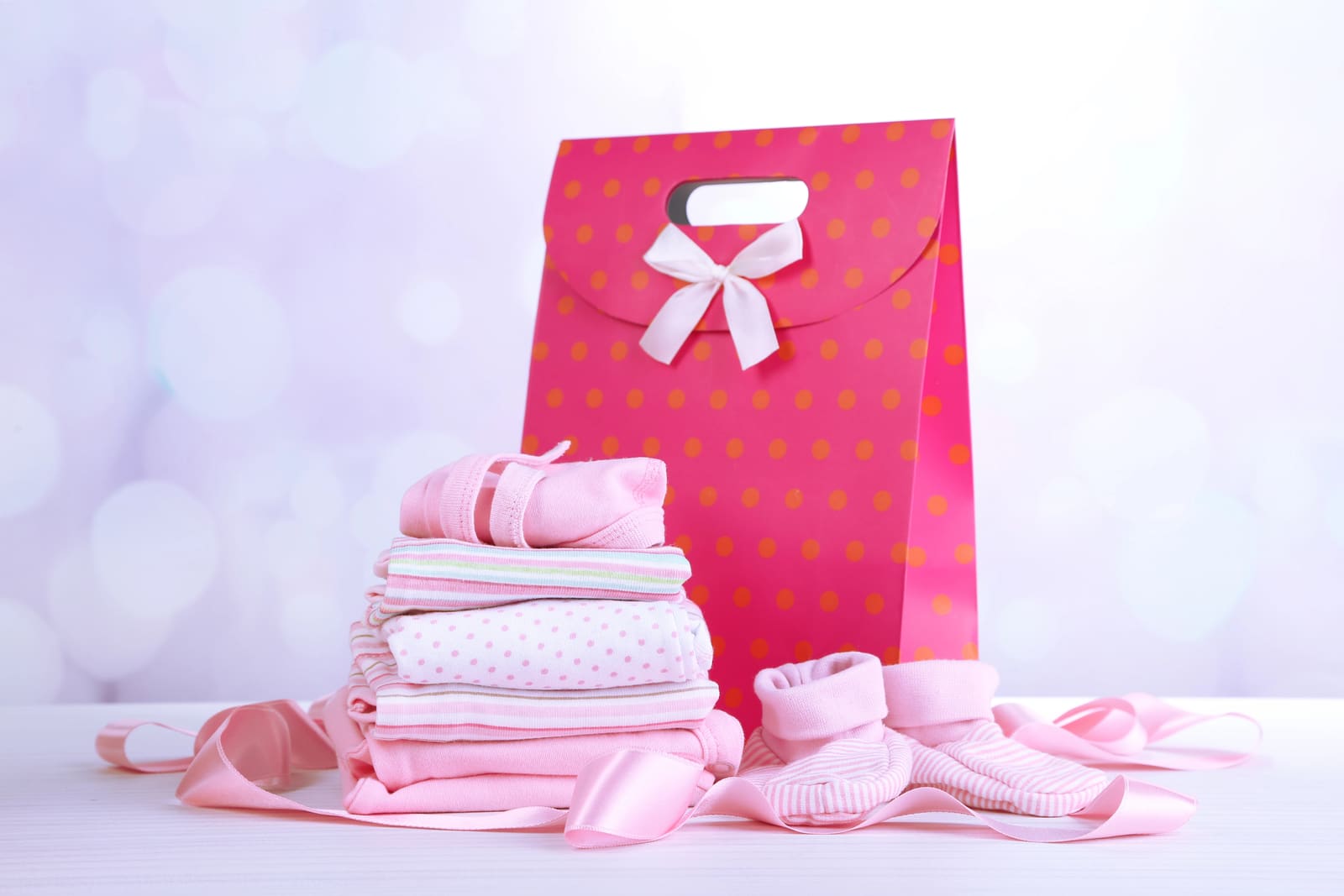 Pink baby clothes and booties beside a pink dotted gift bag with a bow