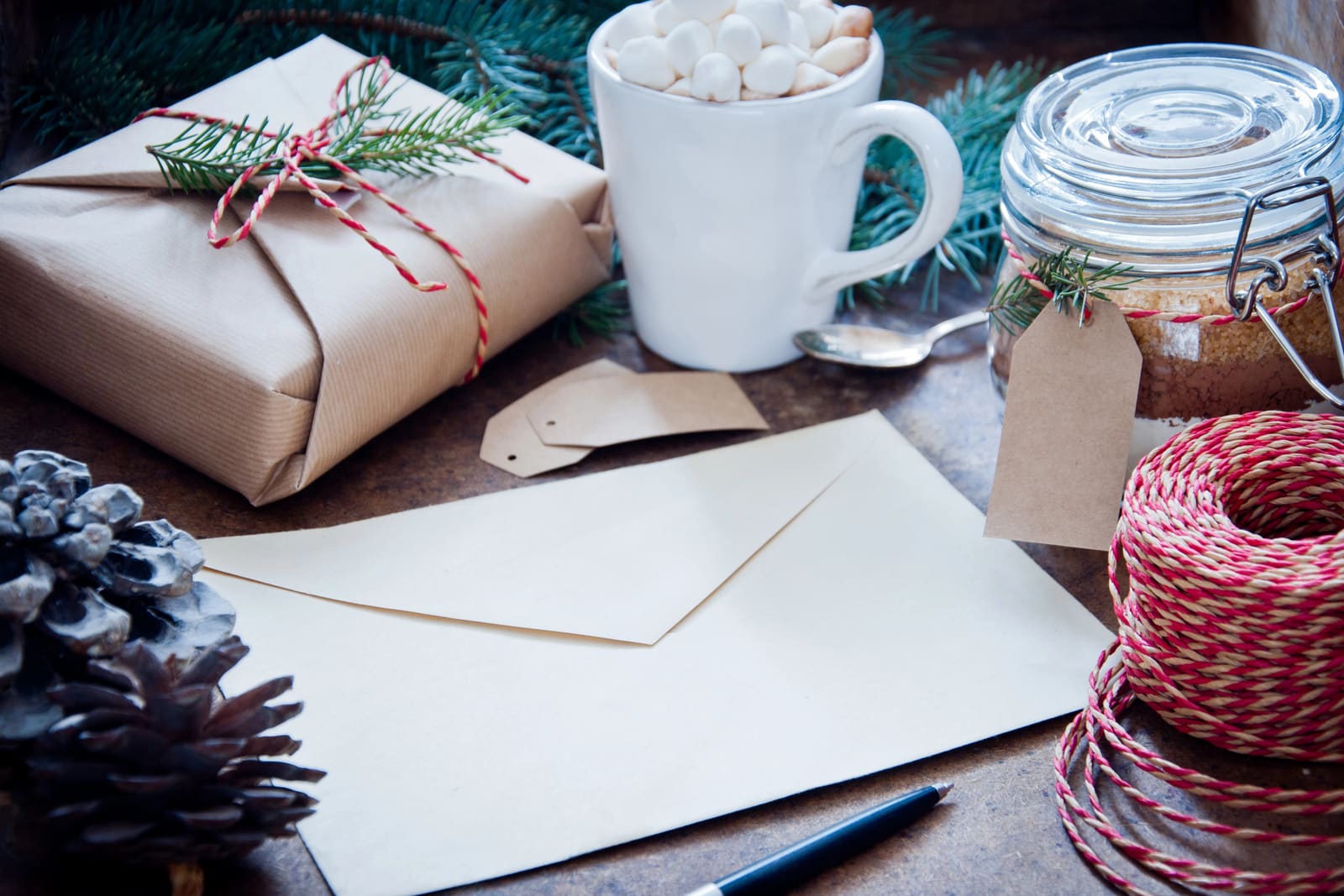 A plain envelope beside a wrapped parcel, red twine and a mug of cocoa