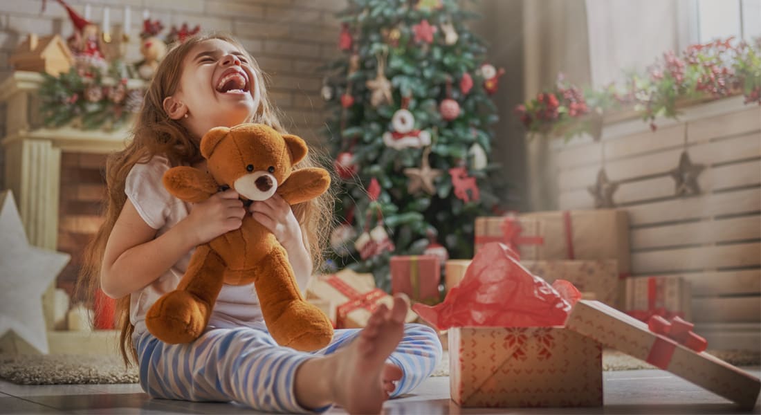 A laughing girl in pyjamas hugs a teddy bear on the floor by a Christmas tree, with open gift boxes beside her