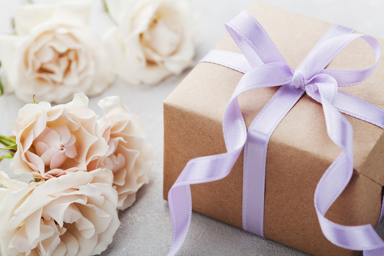 Wedding gift with a purple ribbon and white roses lying next to it.