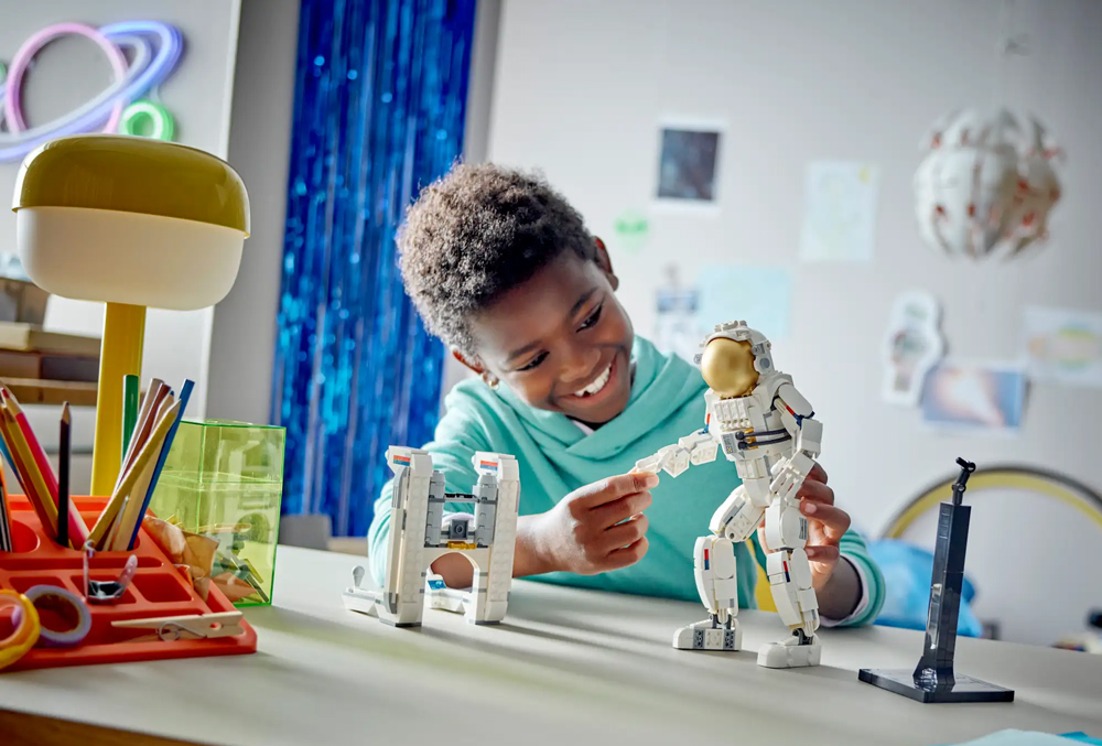 Dziecko w turkusowej bluzie bawiące się kombinezonem mechanicznym astronauty LEGO i podstawką wystawową